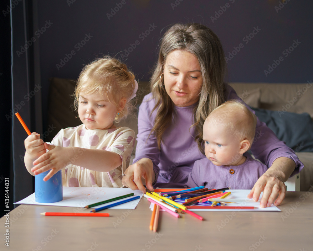 Young loving mother drawing with colored pencils with two adorable little kids at home on dark background, moody day. Happy mom teaching small daughter and baby boy son how to draw. Motherhood concept Young loving mother drawing with colored pencils with two adorable little kids at home on dark background, moody day. Happy mom teaching small daughter and baby boy son how to draw. Motherhood concept