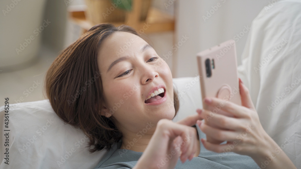 happy asian woman face relaxing using mobile smart phone surfing internet and chat on sofa couch at home. female laying down in living room embarrassed smile when using mobile phone. woman laugh happy happy asian woman face relaxing using mobile smart phone surfing internet and chat on sofa couch at home. female laying down in living room embarrassed smile when using mobile phone. woman laugh happy