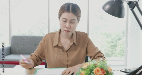 Front view of asian women writing and lectuing note or information on her folder and talking to the camera in her office. Concept online video confernece photo