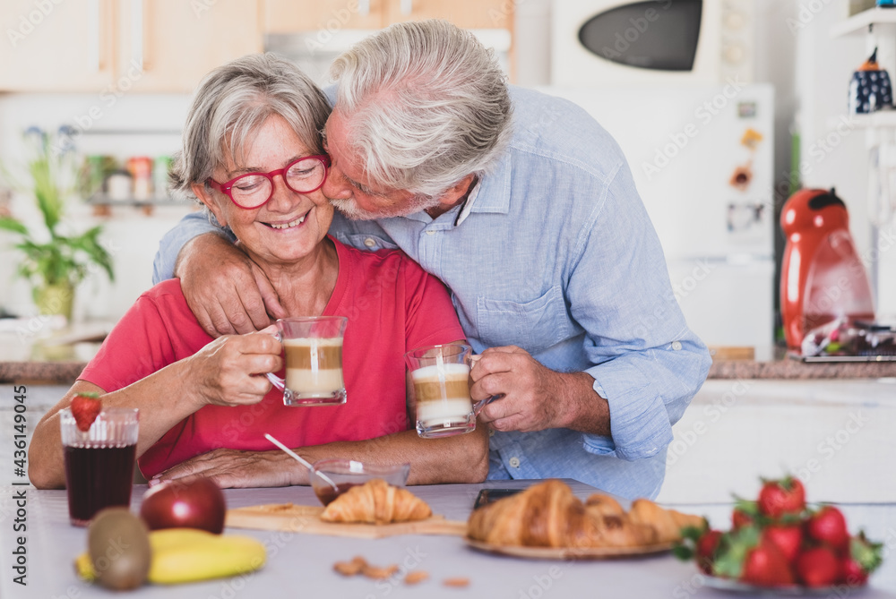 Beautiful senior couple having breakfast at home, starting morning with a kiss. Hot cappuccino in the hands
