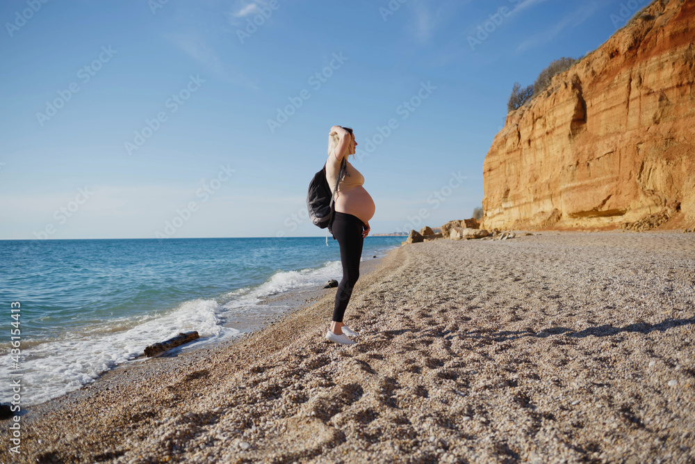 young pregnant woman with a backpack walks along the sea