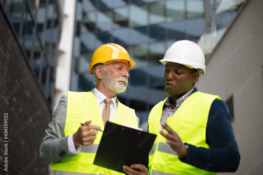 Mature engineer discussing the structure of the building with architects colleague at construction site. Reading document. Mature engineer discussing the structure of the building with architects colleague at construction site. Reading document.