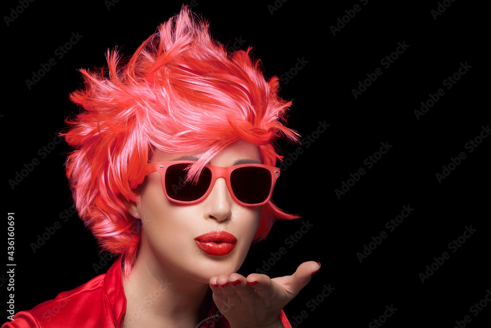 Stylish young woman wearing trendy modern sunglasses and red wig blowing across the palm of her hand sending a kiss