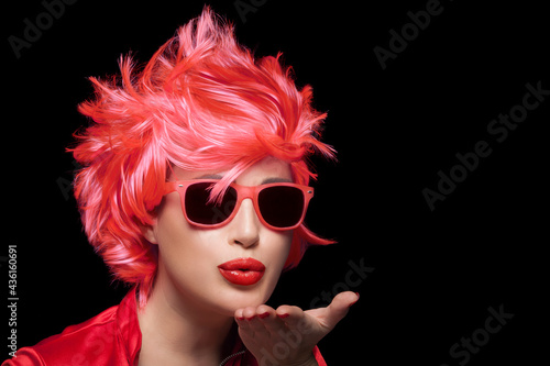 Stylish young woman wearing trendy modern sunglasses and red wig blowing across the palm of her hand sending a kiss photo