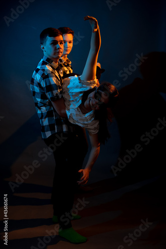 Two guys in a plaid shirt are holding a girl on a dark background in a studio with multicolored light.