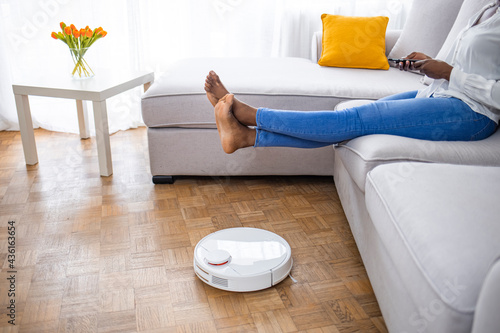 Robot vacuum cleaner cleaning carpet, woman legs rest sitting on sofa at home. Woman lifts feet up when a round robot vacuum cleaner passes to clean the dirty floor.