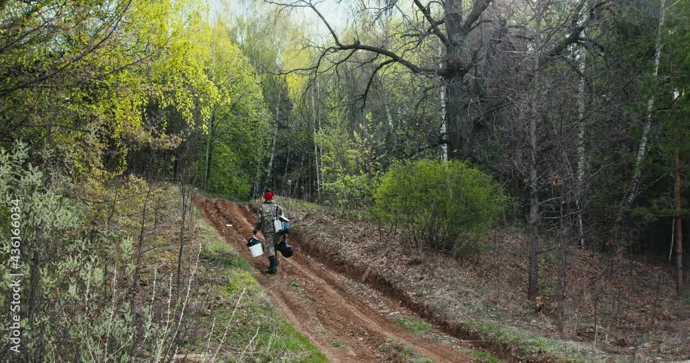 Angler walks with stuff. Fisherman slowly climbs the hill in the spring forest and carries fishing equipment Angler walks with stuff. Fisherman slowly climbs the hill in the spring forest and carries fishing equipment