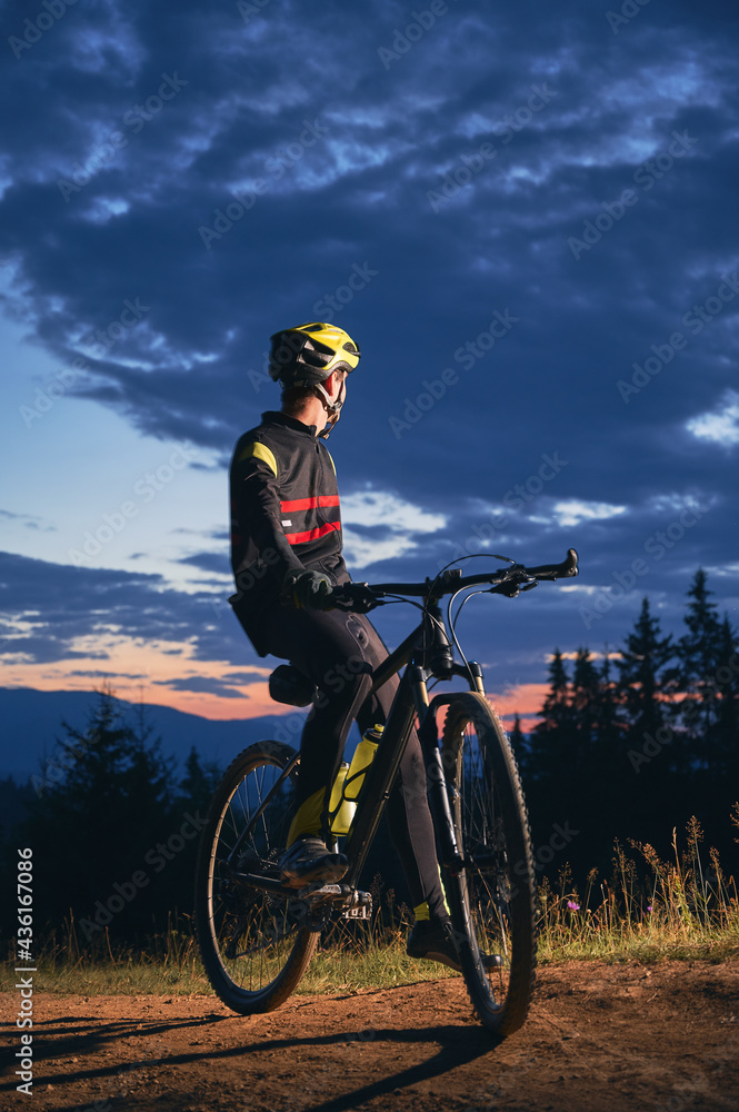 Naklejka premium Young man in cycling suit sitting on bicycle under beautiful night sky. Male bicyclist in safety helmet resting on hillside road under blue cloudy sky while riding bicycle in the evening.