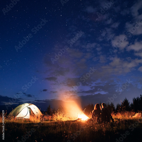 Beautiful view of night starry sky over grassy hill with illuminated camp tent, campfire and hikers. Silhouette of man and woman tourists resting near bonfire. Concept of night camping, relationship.