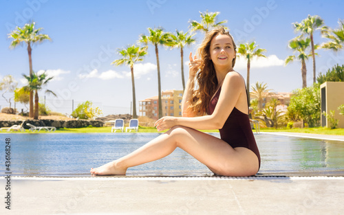 Gorgeous blonde sits near the pool on vacation. A girl with long hair in a single red swimsuit sits on a background of palm trees. Years, sunny day, girl traveling, sunbathing. Summer mood.