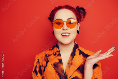 Cute girl on a red background. Red hair and tunnels in the ears, red dress and lips. A woman in red sunglasses and two buns on her head looks at the camera, an air kiss, a place for text