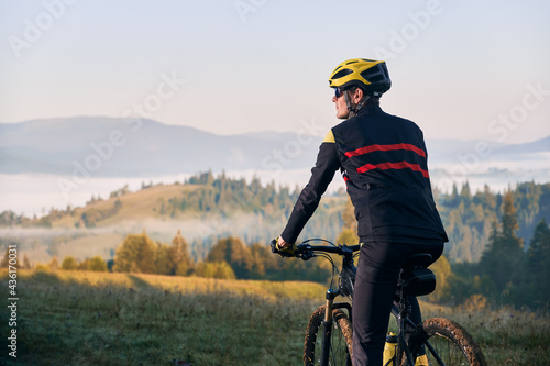 Close up male cyclist in cycling suit riding bike with hills on background. Man bicyclist wearing safety helmet and glasses while enjoying bicycle ride in mountains in the morning. Back view