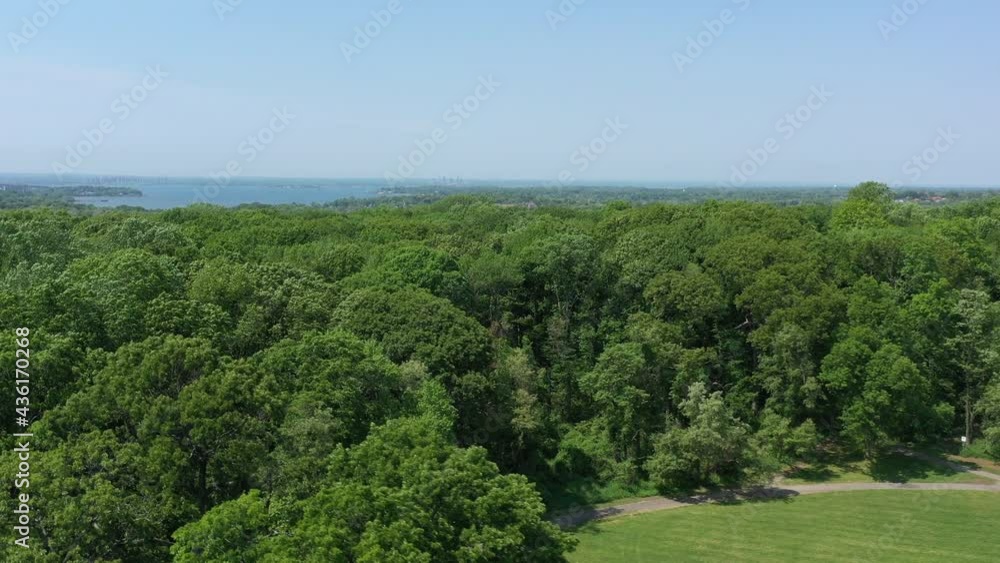 An aerial view above green tree tops in a park on a sunny day. The drone camera, focused on a bay in the far distance, dolly in over the trees and an empty baseball field below. An aerial view above green tree tops in a park on a sunny day. The drone camera, focused on a bay in the far distance, dolly in over the trees and an empty baseball field below.