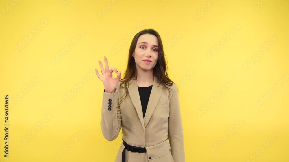 Positive female office worker in suit stands on yellow background with smile on face and shows OK gesture with fingers. Positive female office worker in suit stands on yellow background with smile on face and shows OK gesture with fingers.