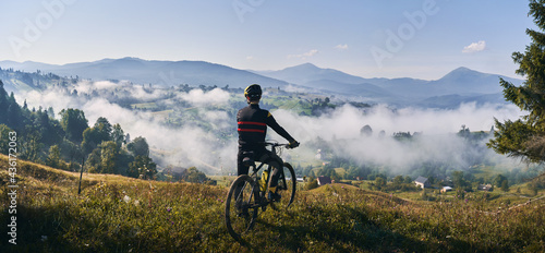 Man riding bicycle on grassy hill and looking at beautiful misty mountains. Male bicyclist enjoying panoramic view of majestic mountains during bicycle ride. Concept of sport, bicycling and nature. photo
