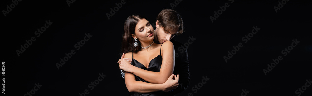 man in black blazer and white shirt kissing shoulder of elegant woman in silk dress and jewelry isolated on black, banner