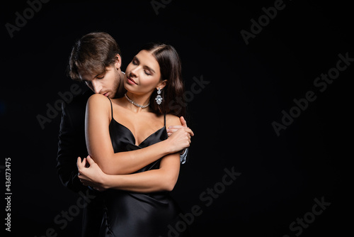 tender man gently kissing shoulder of woman in silk dress and jewelry isolated on black © LIGHTFIELD STUDIOS