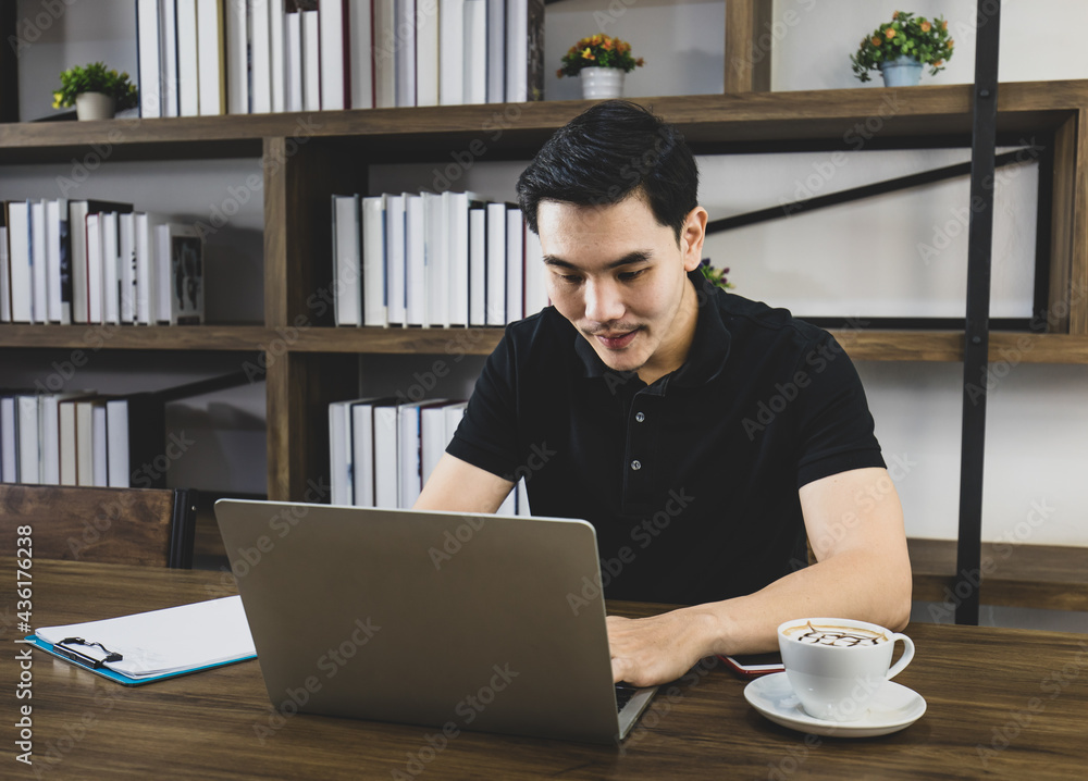 Handsome Asian man in casual black shirt sitting at ease on brown table with cup of morning coffee in reading room and enjoy online business working on laptop with memo paper Handsome Asian man in casual black shirt sitting at ease on brown table with cup of morning coffee in reading room and enjoy online business working on laptop with memo paper