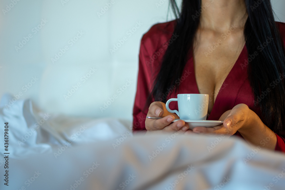 A young female, wearing a red nightdress holding a cup of coffee in her hands in bed on a white blanket. Concept of a relaxed morning in bed.