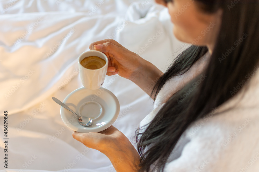 A young female, wearing a bathrobe, holding a cup of coffee in her hands in bed on a white blanket. Concept of a relaxed morning in bed.