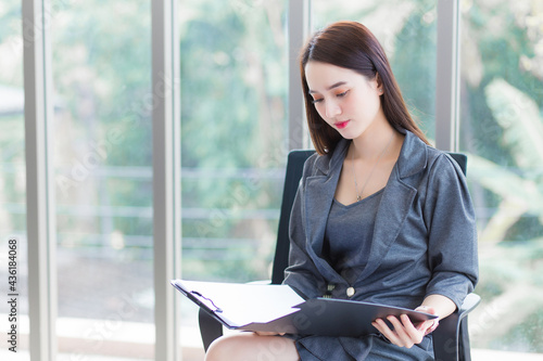 Asian professional working female who wears grey dress sits and looks at documents in a office chair while she works seriously.