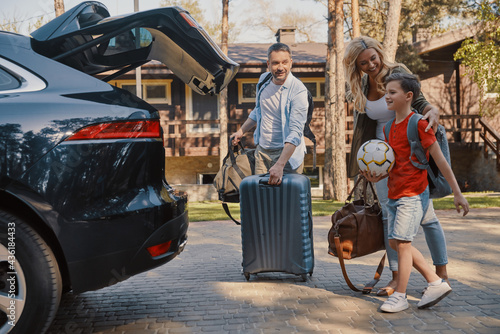 Happy young family with little packing stuff into the car and smiling while standing near house