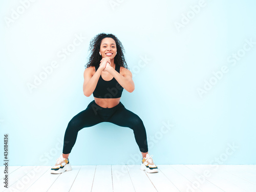 Fitness smiling black woman in sports clothing with afro curls hairstyle.She doing squats. Young beautiful model with perfect tanned body.Female in studio near light blue wall.Cheerful and happy