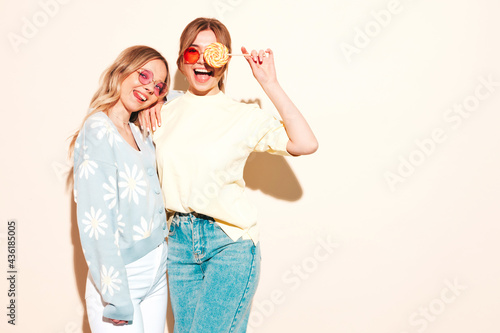 Two young beautiful blond smiling hipster female in trendy summer clothes. Sexy carefree women posing in studio. Trendy and positive models having fun with candy sweet lollipop © halayalex