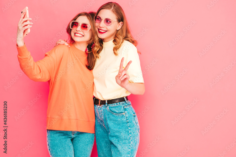 Two young beautiful smiling hipster female in trendy summer hipster clothes. Sexy carefree women posing near pink wall in studio. Trendy and positive models having fun. Taking selfie photos Two young beautiful smiling hipster female in trendy summer hipster clothes. Sexy carefree women posing near pink wall in studio. Trendy and positive models having fun. Taking selfie photos