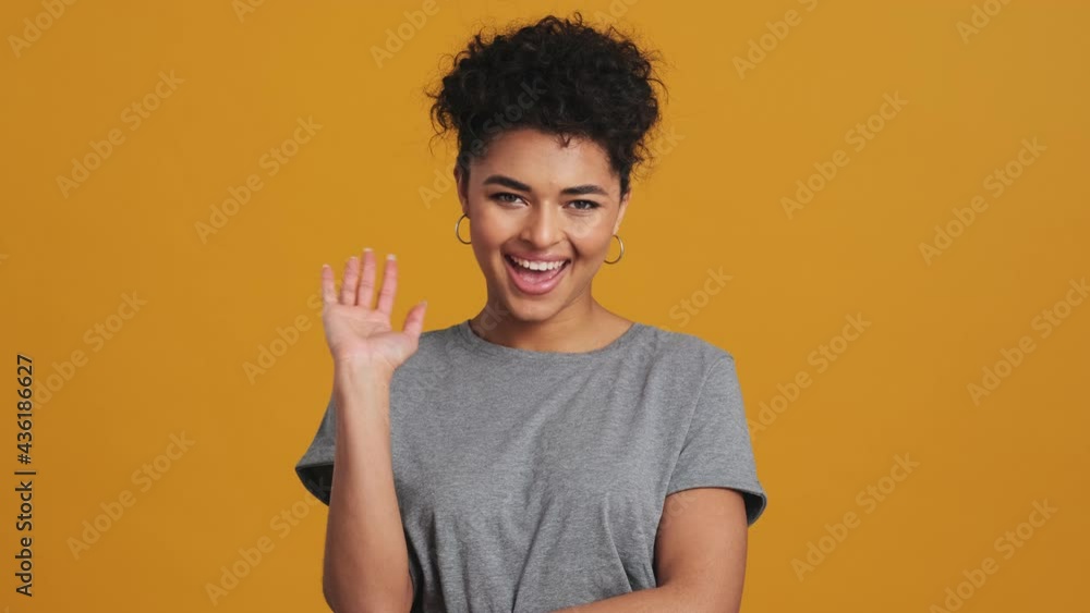 A positive american woman wearing gray basic T-shirt is doing hello gesture isolated over yellow wall in the studio A positive american woman wearing gray basic T-shirt is doing hello gesture isolated over yellow wall in the studio