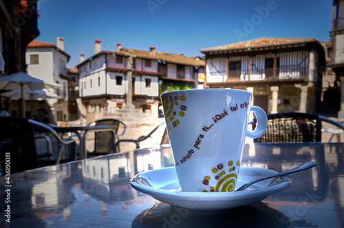 Close-Up Coffee Cup On Metal Table On The Terrace Of A Bar With Townhouses Out Of Focus In The Background.  On The Cup You Can Read..."one minute for you...how about this one?"