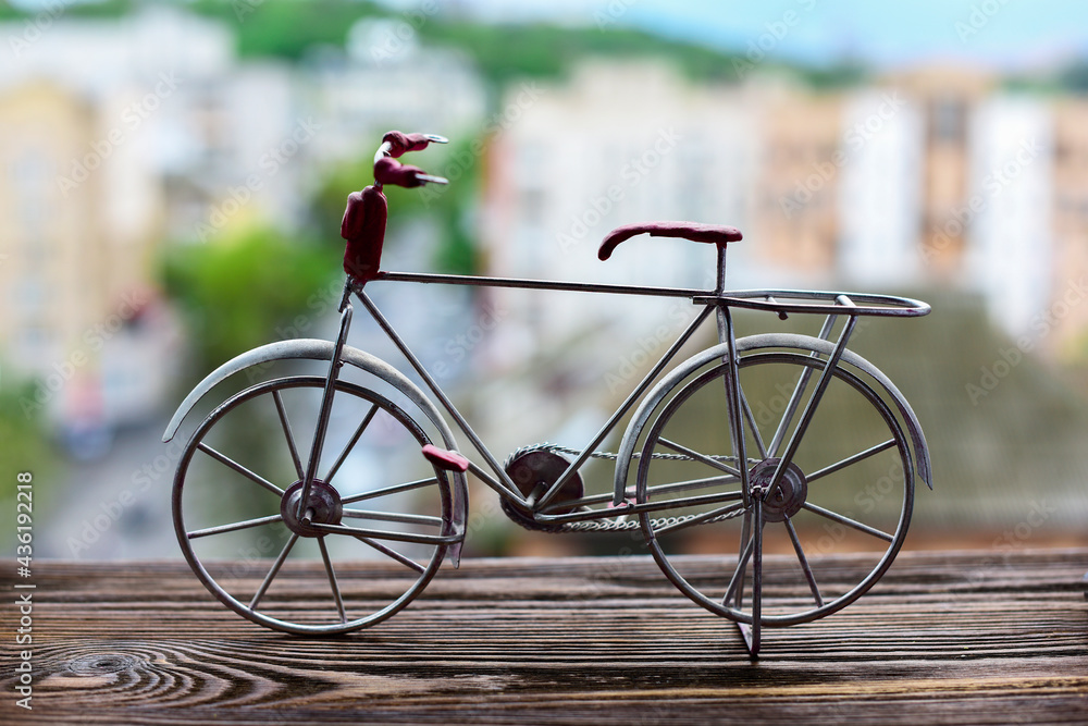 Close-up of a toy souvenir metal bicycle on a wooden windowsill against a blurry background of a city summer landscape. World Bicycle Day concept. Sport, Recreation and Health.
