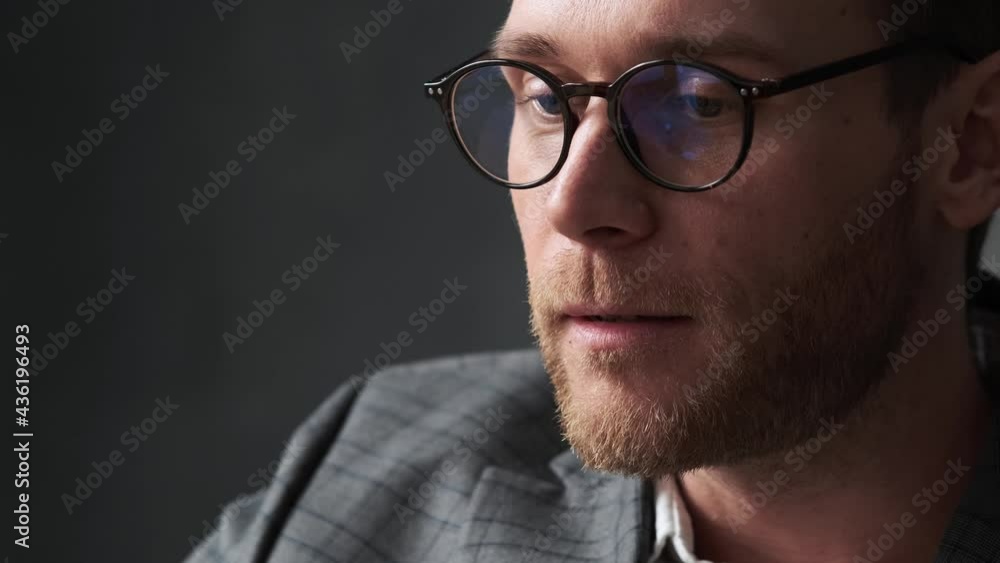 A close-up view of a pensive man in an office suit and glasses looking forward and biting his lips in a gray office A close-up view of a pensive man in an office suit and glasses looking forward and biting his lips in a gray office