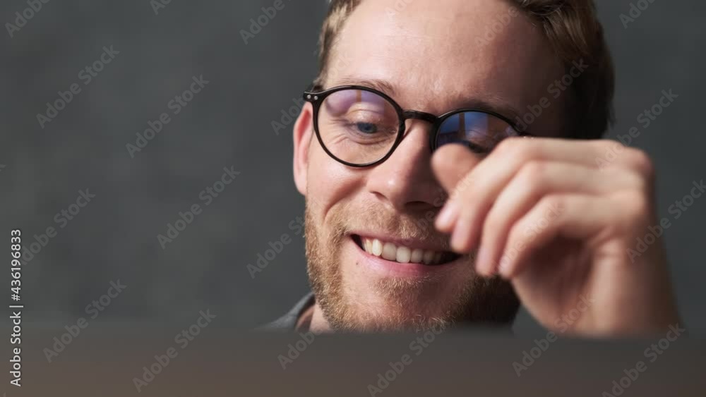 A close-up view of a smiling man wearing glasses and putting on wireless headphones looking at a laptop in a gray office A close-up view of a smiling man wearing glasses and putting on wireless headphones looking at a laptop in a gray office