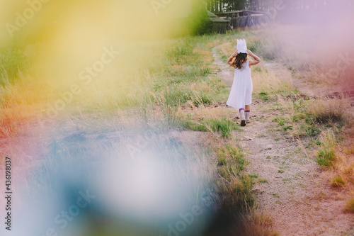 Summertime and children outdoor games concept. Little girl in white dress holding handmade crown on head moving away across summer field. Blurred frontground. Rear view. Soft focus. Horizontal shot photo