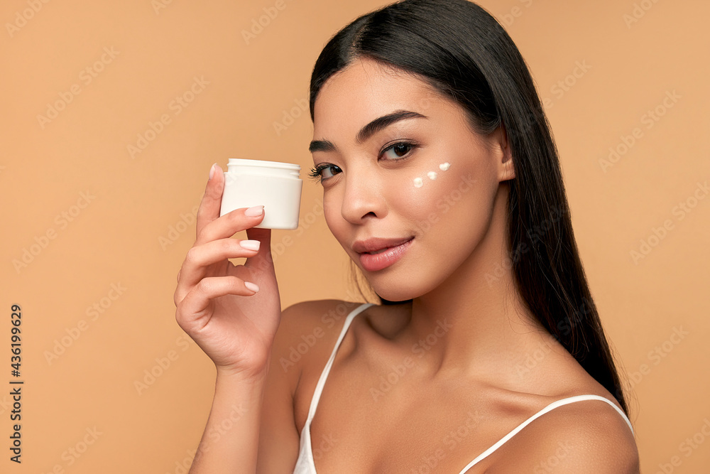 Young Asian woman with clean radiant skin uses a moisturizing face cream on a beige background. Spa care, facial skin care, beauty cosmetology. Young Asian woman with clean radiant skin uses a moisturizing face cream on a beige background. Spa care, facial skin care, beauty cosmetology.