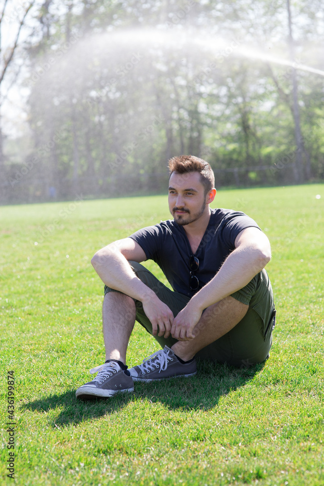 Junger Mann mit schwarzen Haaren und einem Bart sitzt auf dem Boden und schaut zur Seite. Er geniesst das Leben und entspannt sich einfach. Hinter Ihm spritzt das Wasser, begießt den Boden. Junger Mann mit schwarzen Haaren und einem Bart sitzt auf dem Boden und schaut zur Seite. Er geniesst das Leben und entspannt sich einfach. Hinter Ihm spritzt das Wasser, begießt den Boden.