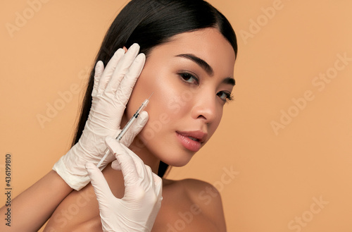 Young Asian woman with clean radiant skin gets botox injections for contour tightening, lip augmentation on a beige background. Spa care, facial skin care, beauty cosmetology. photo