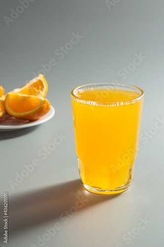 Tablou pe pânză Orange carbonated drink in a glass and orange slices in a plate in the background