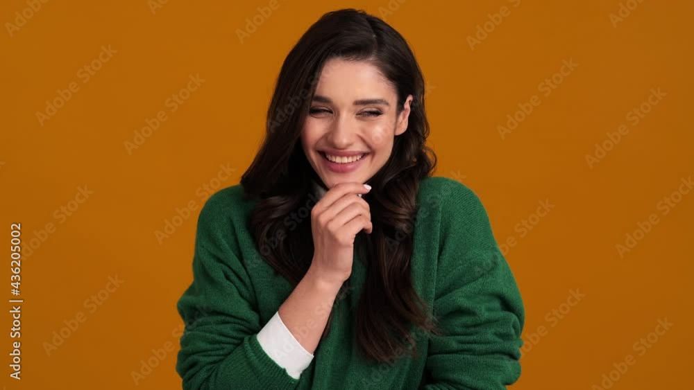 A positive young woman in a green sweater and shirt looking to the sides and laughing while standing in an orange studio A positive young woman in a green sweater and shirt looking to the sides and laughing while standing in an orange studio