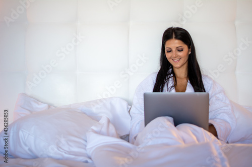 Beautiful young woman sitting in bed using laptop in a modern room. Caucasian model wearing a bathrobe relaxing with computer indoors, surfing the internet, shopping online and home office 
