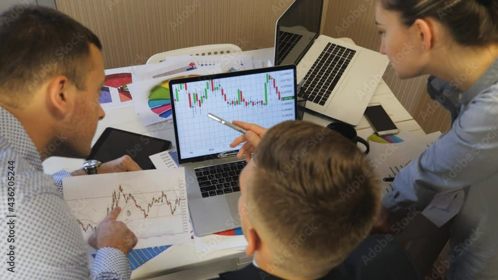 Professional managers comparing sales result graphs on papers with chart on computer screen at office. Group of young colleagues analyzing financial reports during business meeting indoor. Top view Professional managers comparing sales result graphs on papers with chart on computer screen at office. Group of young colleagues analyzing financial reports during business meeting indoor. Top view