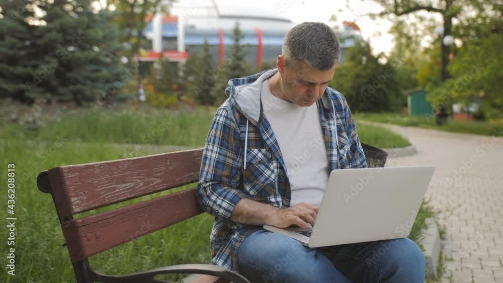 A male freelancer sits on a bench in a park and works on a laptop. A male freelancer sits on a bench in a park and works on a laptop.