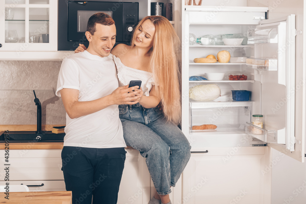 Ordering food through the online store. A couple of lovers in the kitchen at home near the refrigerator. Man and Woman are shopping online through the phone. Smart refrigerator. Ordering food through the online store. A couple of lovers in the kitchen at home near the refrigerator. Man and Woman are shopping online through the phone. Smart refrigerator.