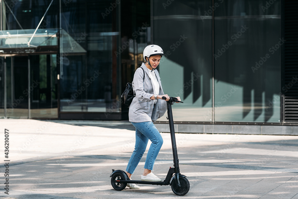 Young businesswoman with a safety helmet on her head driving electrical scooter near an office building Young businesswoman with a safety helmet on her head driving electrical scooter near an office building