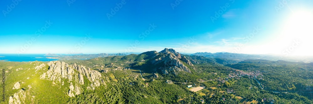 View from above, stunning aerial view of San Pantaleo village surrounded by some granite mountains, green vegetation and the mediterranean sea in the distance. Sardinia, Italy.