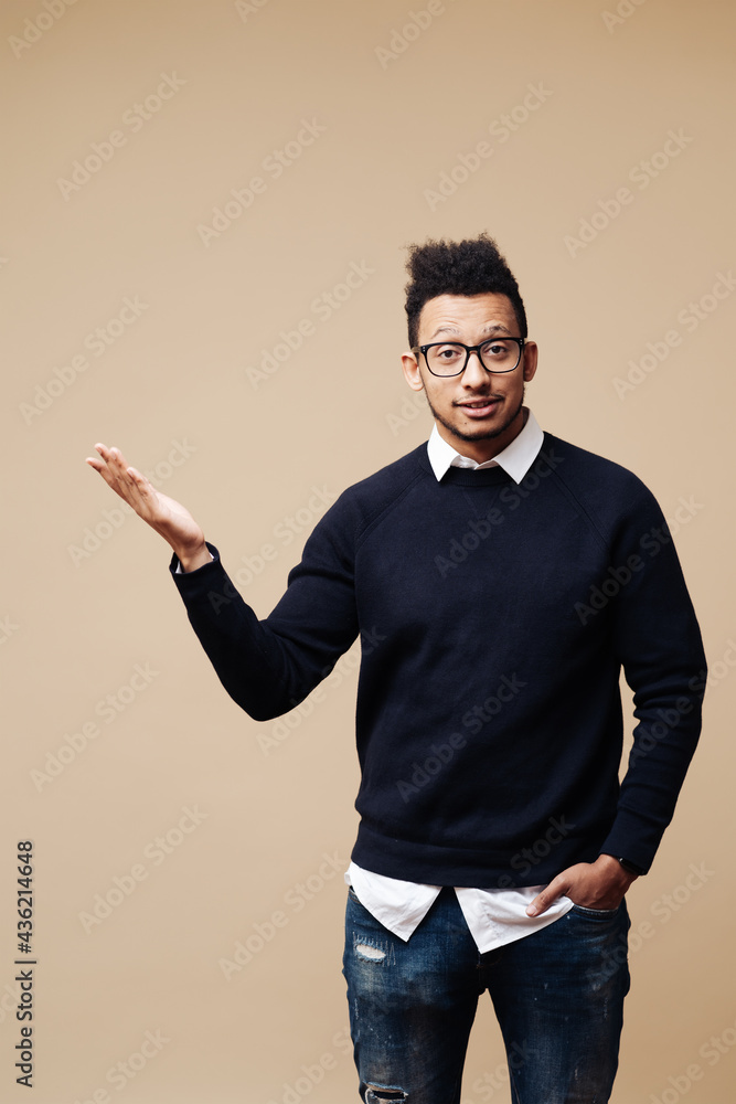 Portrait of handsome young Afro man wearing glasses smiling and standing with open hand gesture isolated on beige background with copy space Portrait of handsome young Afro man wearing glasses smiling and standing with open hand gesture isolated on beige background with copy space