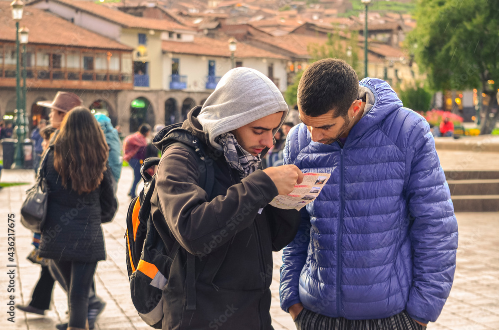 Two young male tourists holding and checking a map in travel destination city. Main city square (barrack square) of Cusco in Peru. Friends walking tour in cold morning