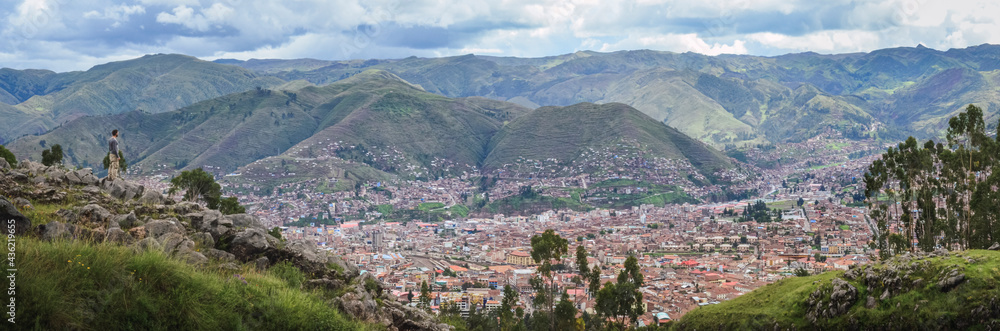 Panoramic view of a young man on top of a hill contemplating Cusco city landscape from the Sacsayhuaman inca archaeological site valley and ancient fortress. Peru, South America