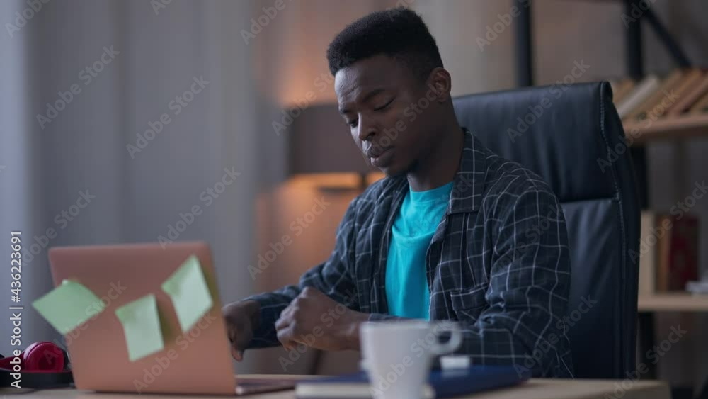 Middle shot of sad African American young man stretching back spasm sitting at table with laptop. Portrait of unwell millennial having chronic problems working in home office. Sedentary lifestyle Middle shot of sad African American young man stretching back spasm sitting at table with laptop. Portrait of unwell millennial having chronic problems working in home office. Sedentary lifestyle